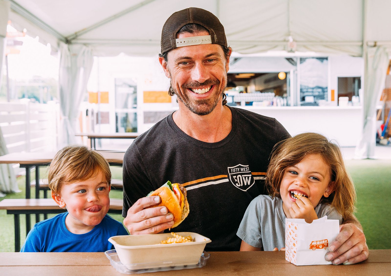 A dad and his two kids enjoying Fifty West burgers and fries at a picnic table